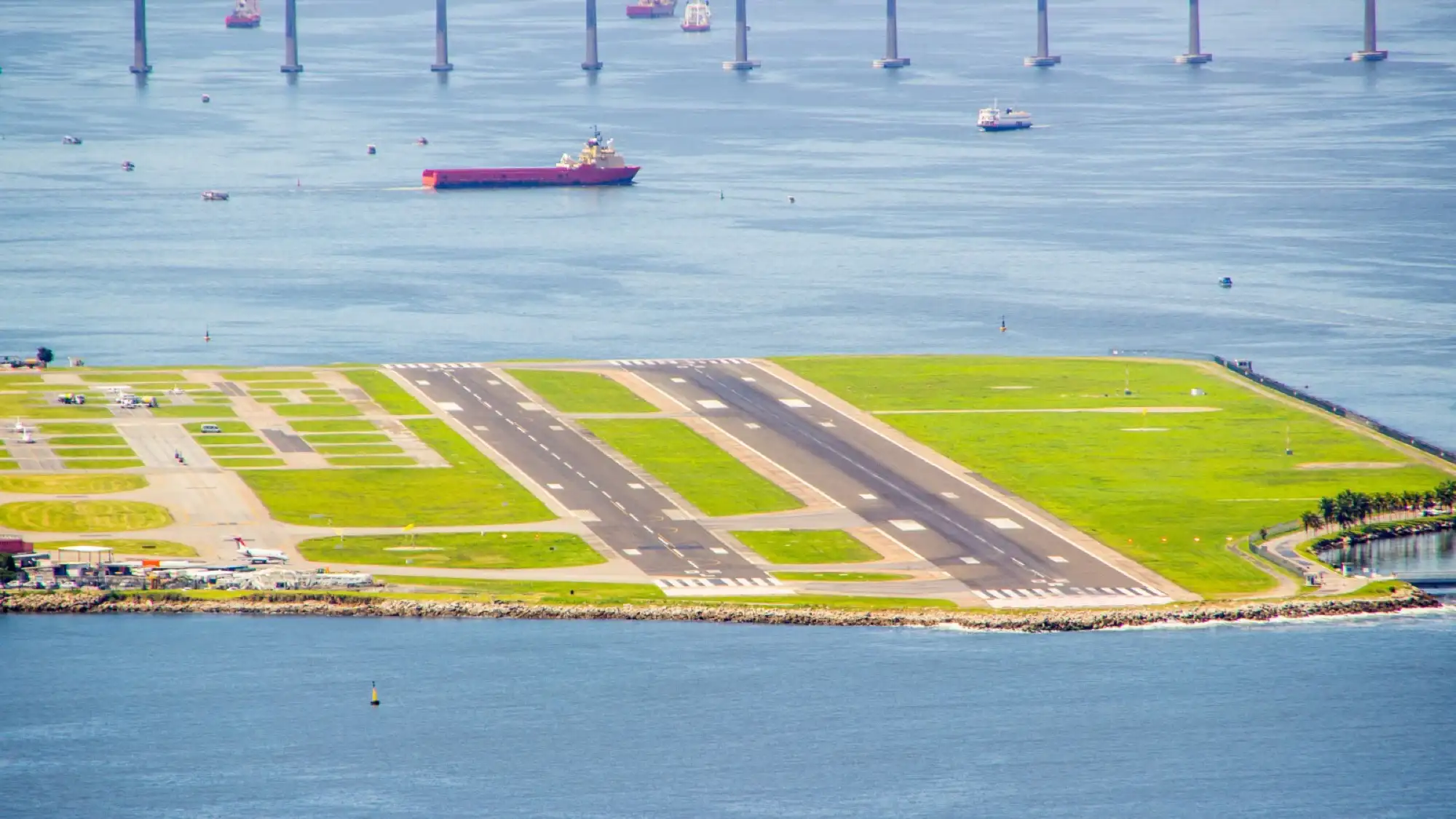 Close-up aerial view of a runway at Rio de Janeiro's Santos Dumont airport, with water, ships, and a long bridge in the background.