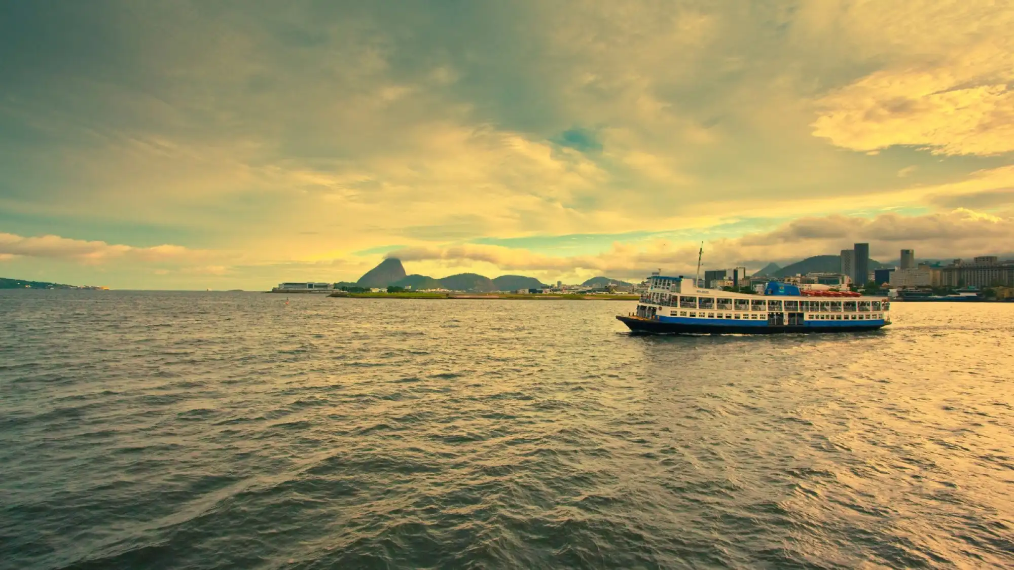 A ferry boat sailing across Guanabara Bay in Rio de Janeiro at sunset, with the city skyline and Sugarloaf Mountain in the background.
