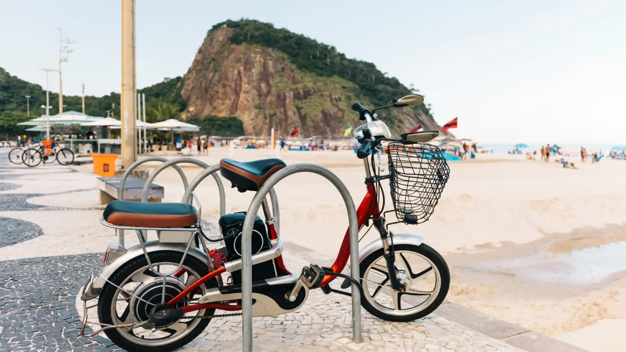 A red electric bike parked at a beachside rack in Rio de Janeiro, with Sugarloaf Mountain and people on the sand in the background.