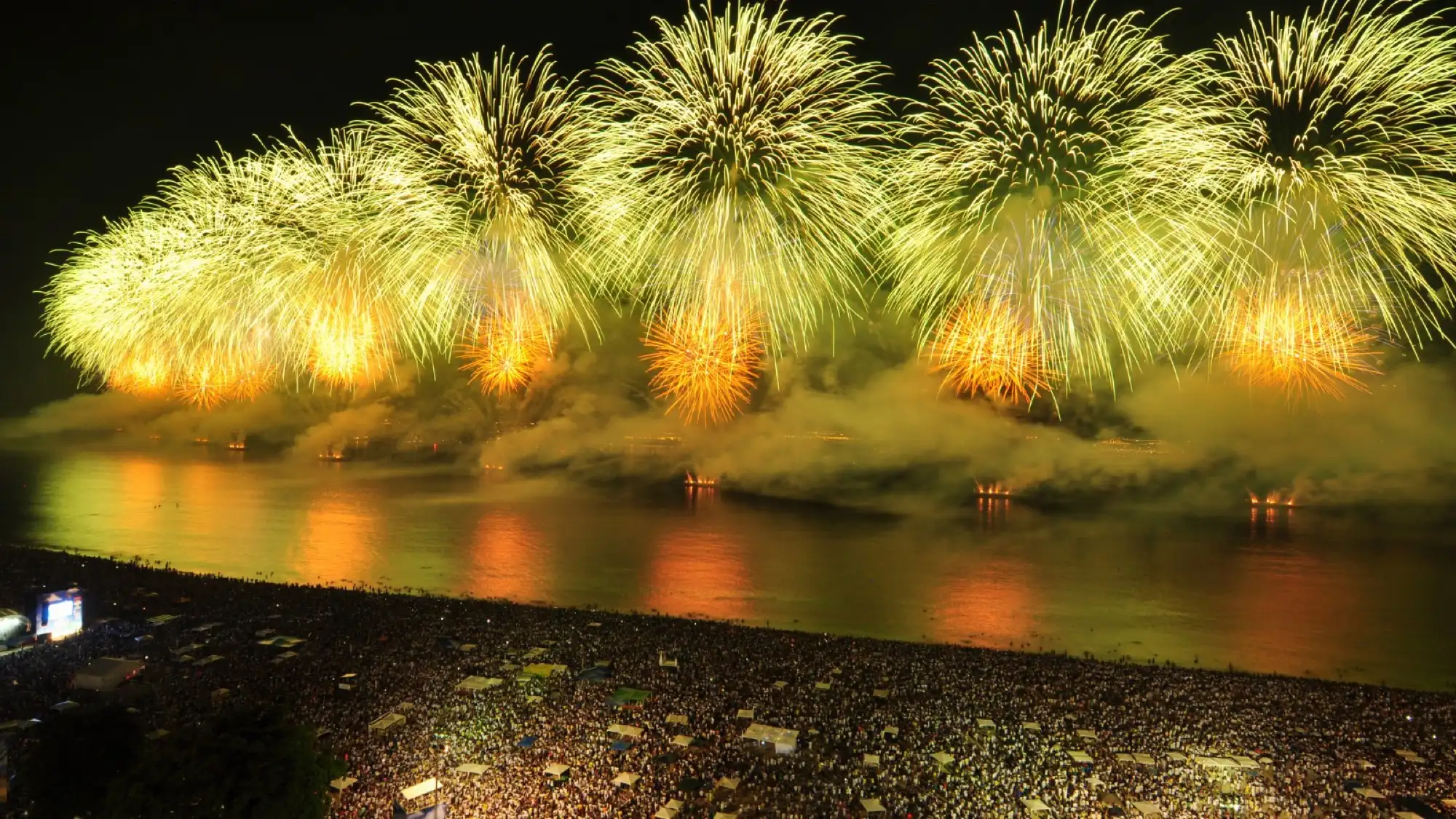 Spectacular fireworks display over Copacabana Beach in Rio de Janeiro, illuminating the night sky above a massive crowd.