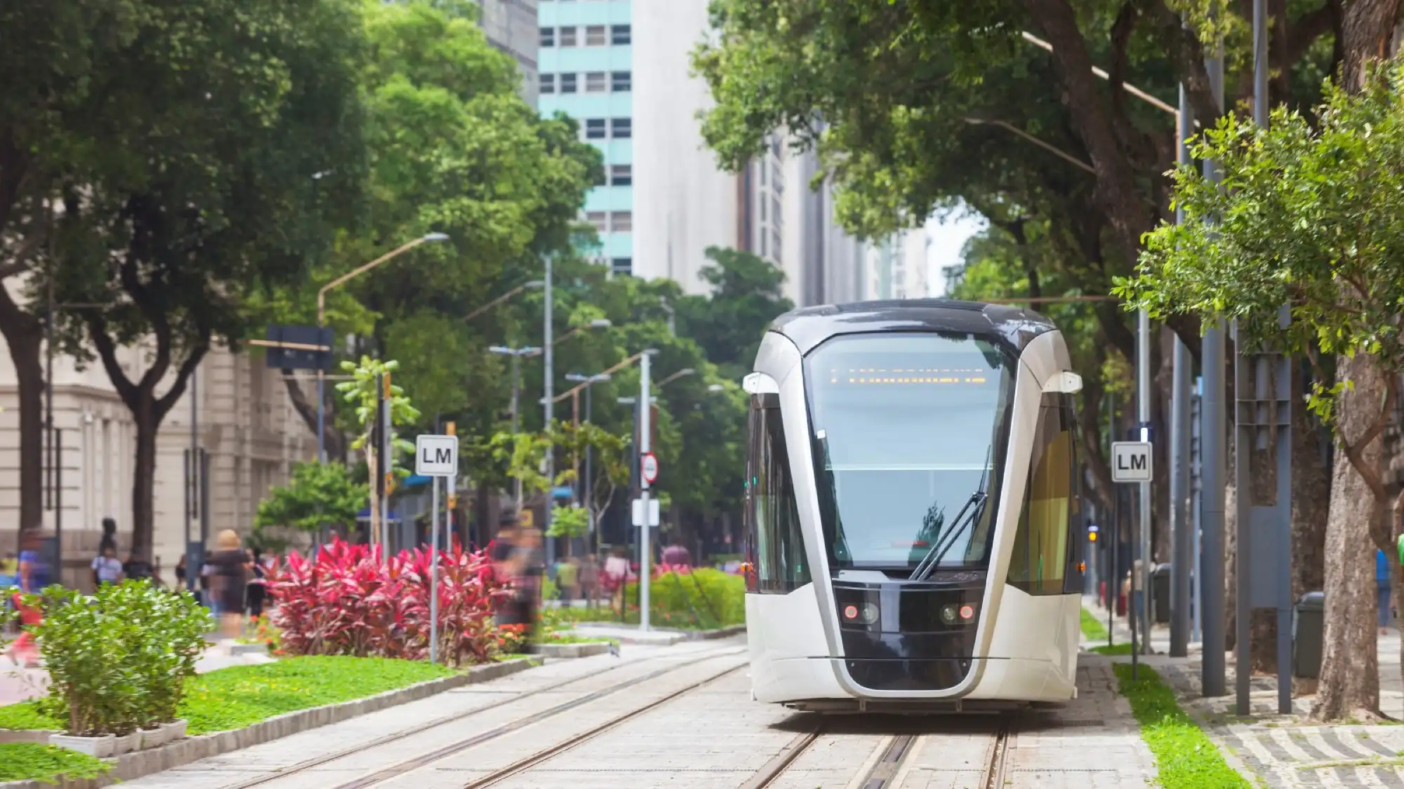 A sleek modern vlt tram moving through a tree-lined street in Rio de Janeiro, surrounded by greenery and urban buildings.