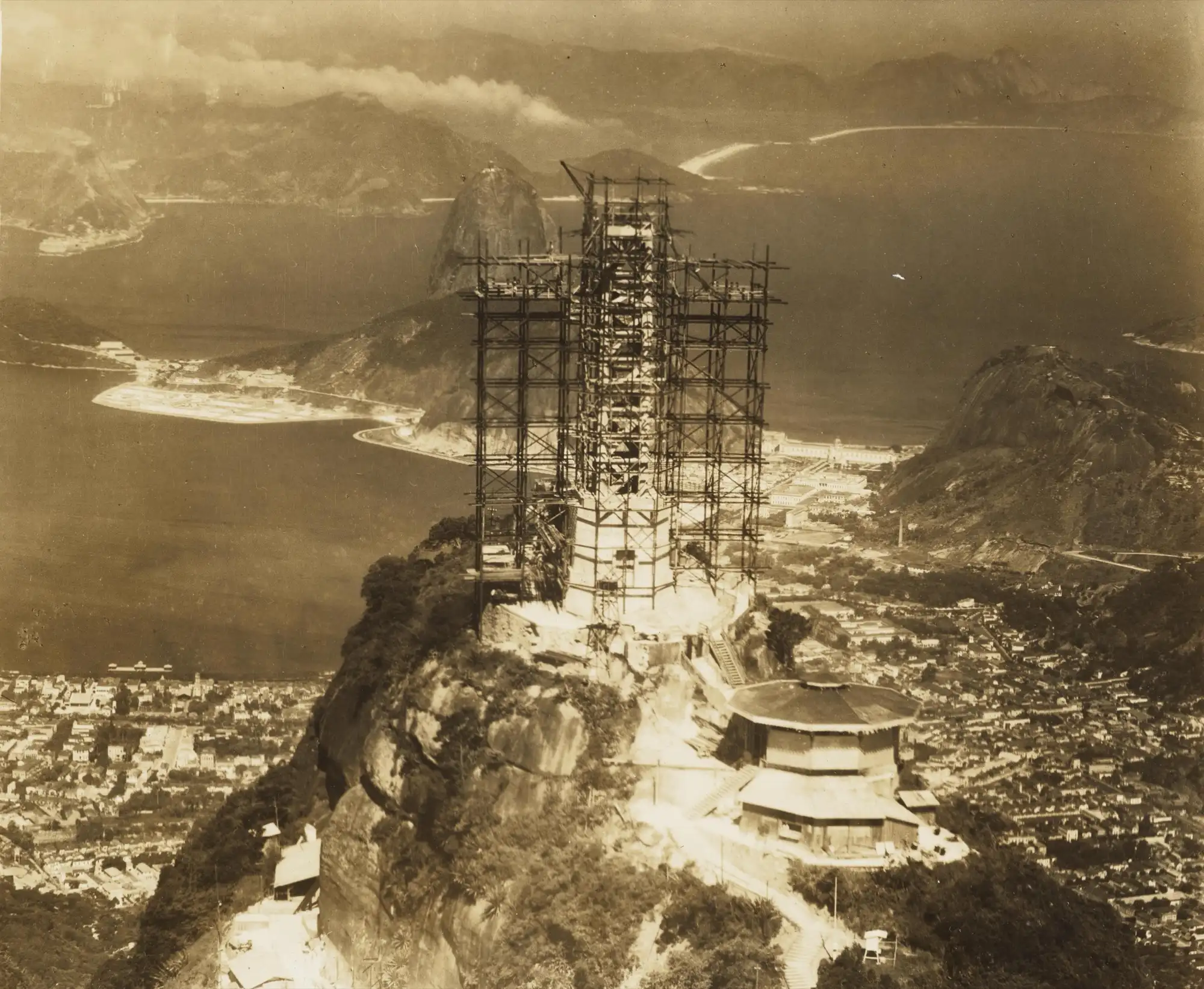 The statue under scaffolding on the summit of Corcovado during building, with Sugarloaf and Guanabara Bay behind
