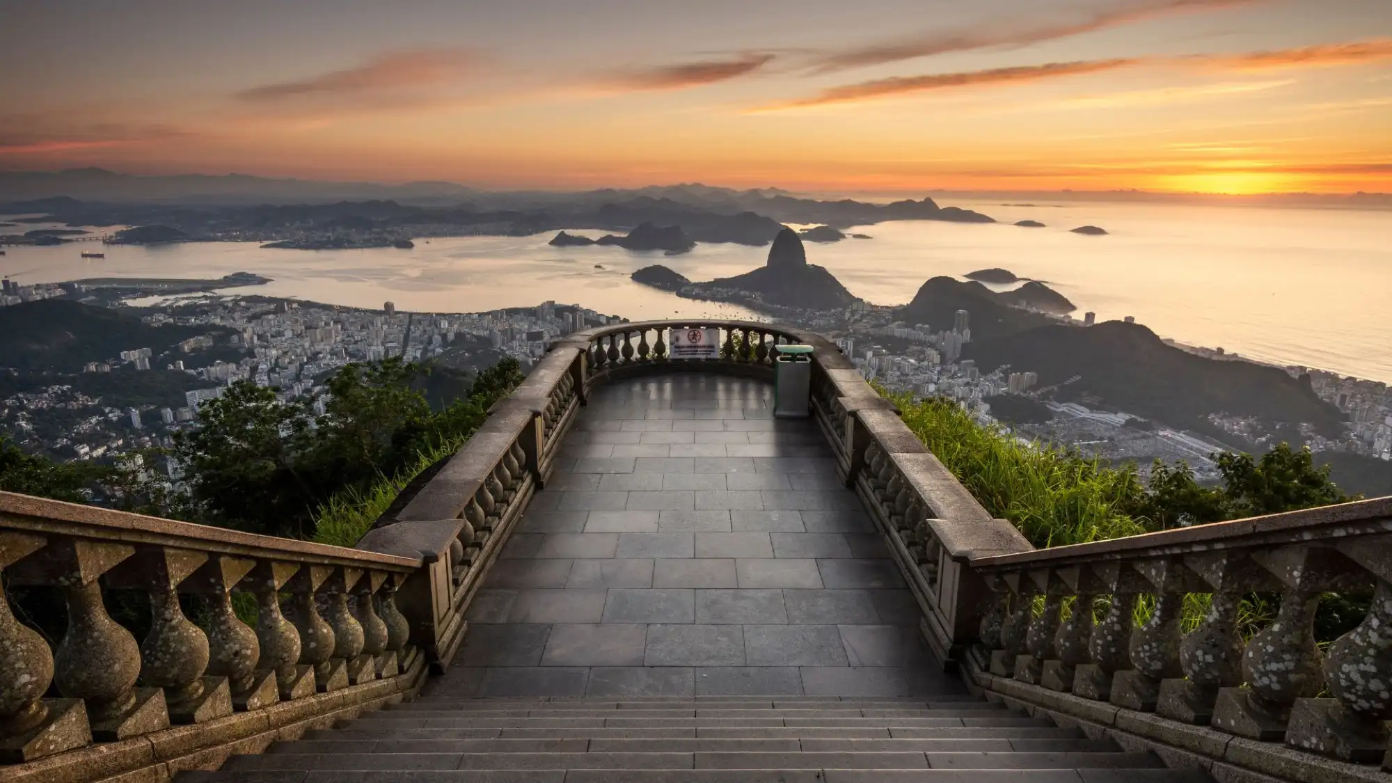 Stone-paved overlook in Tijuca Forest, offering a panoramic view of Rio de Janeiro and the coastline at sunset, with warm orange tones in the sky.