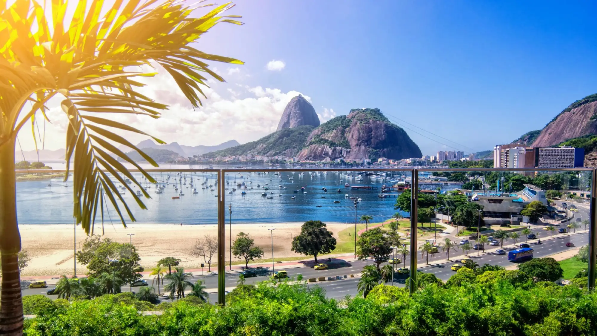 Scenic view of Sugarloaf Mountain and Guanabara Bay in Rio de Janeiro, with palm trees, boats, and a sunny blue sky.