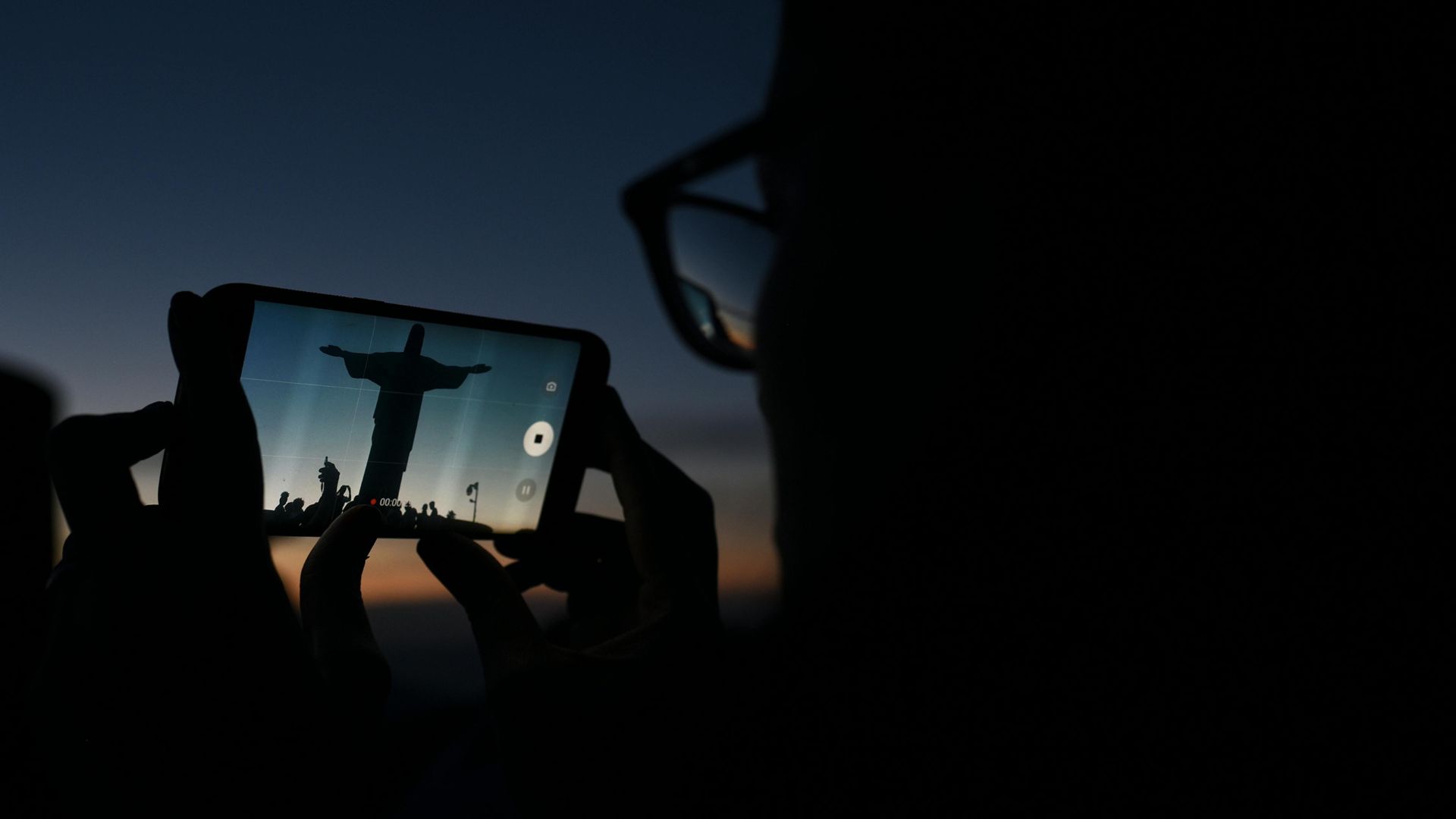 Silhouette of Christ the Redeemer statue captured on a smartphone at dusk, with a person holding the phone and the sky darkening in the background.