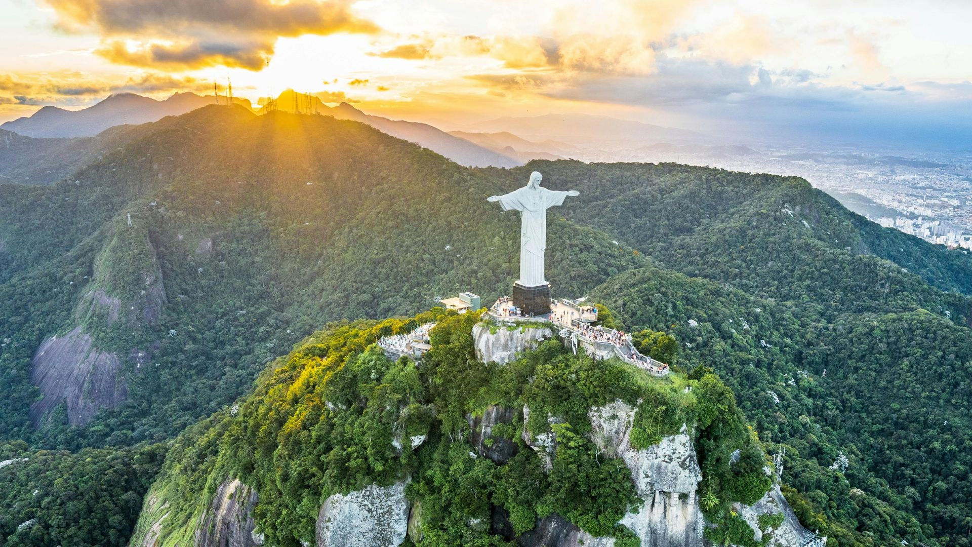 Christ the Redeemer statue at sunset on a mountain in Rio de Janeiro, surrounded by lush green forest with mountains in the background.