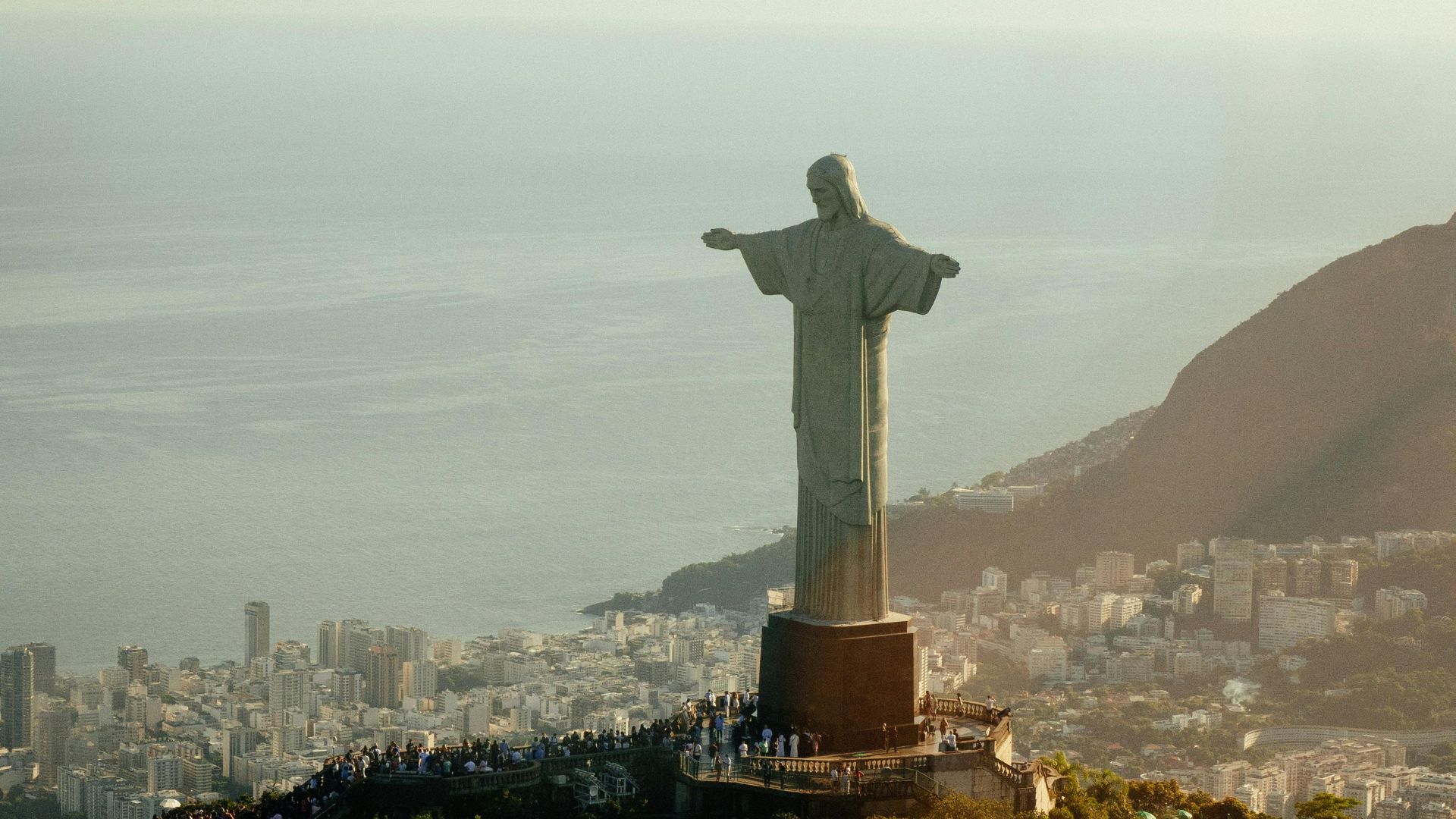 Aerial view of the Christ the Redeemer statue in Rio de Janeiro, overlooking the city and ocean with buildings and mountains in the background.