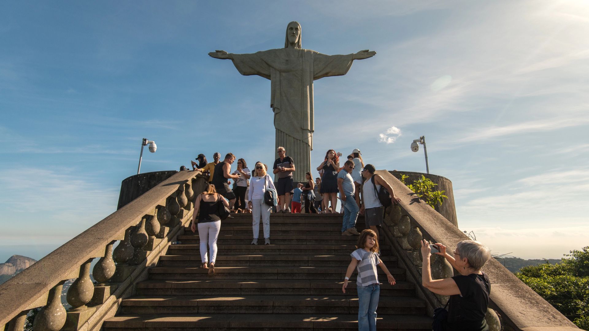 Visitors standing on the steps in front of the Christ the Redeemer statue in Rio de Janeiro, with clear skies in the background.