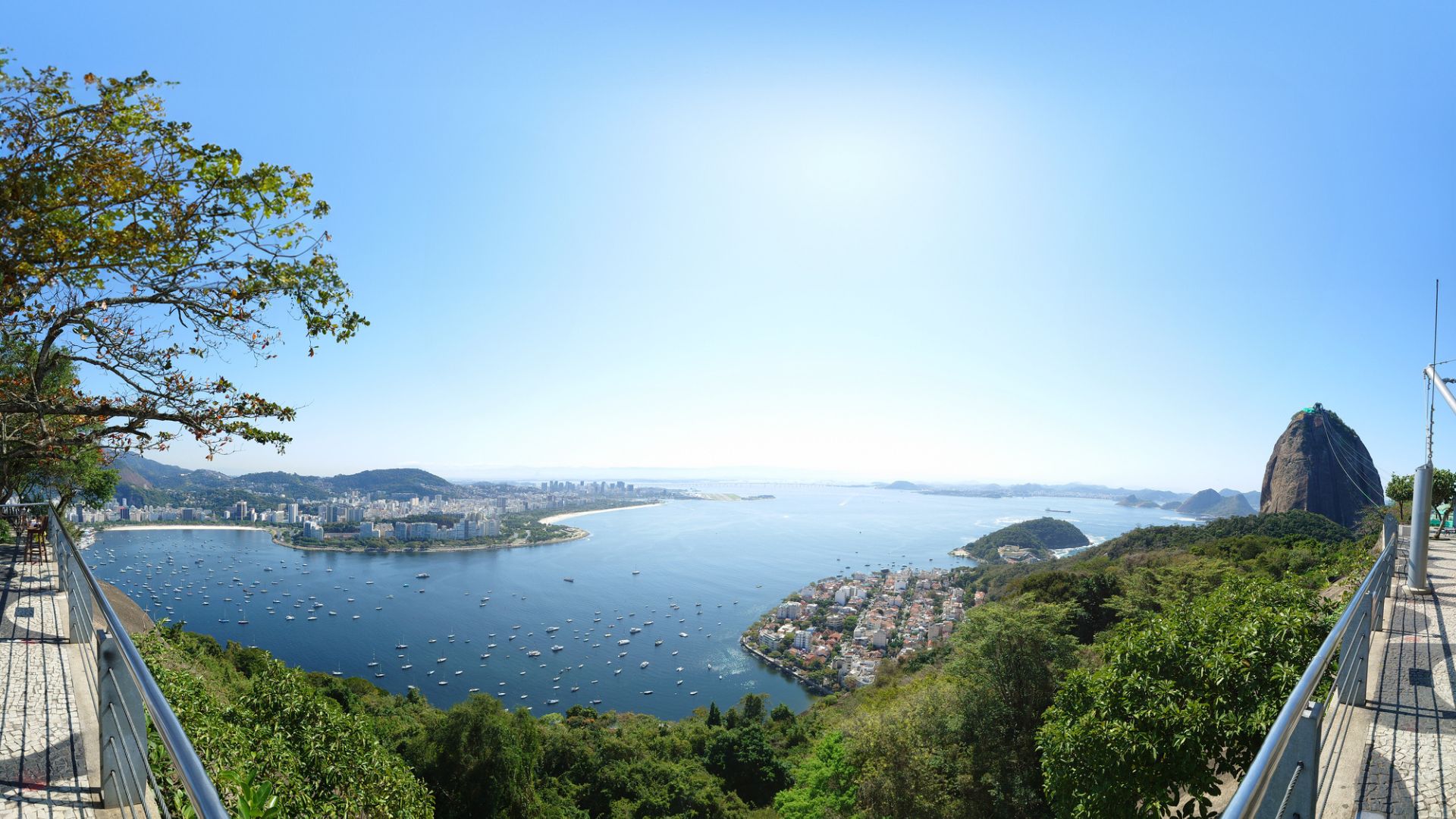 A panoramic view of Rio de Janeiro's coastline, showing the city, boats in the bay, and the iconic Sugarloaf Mountain under a clear blue sky.